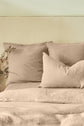 A neatly made bed with beige linen bedding, two large pillows, a smaller pillow, and a sprig of dried leaves resting beside the pillows against a light wall.