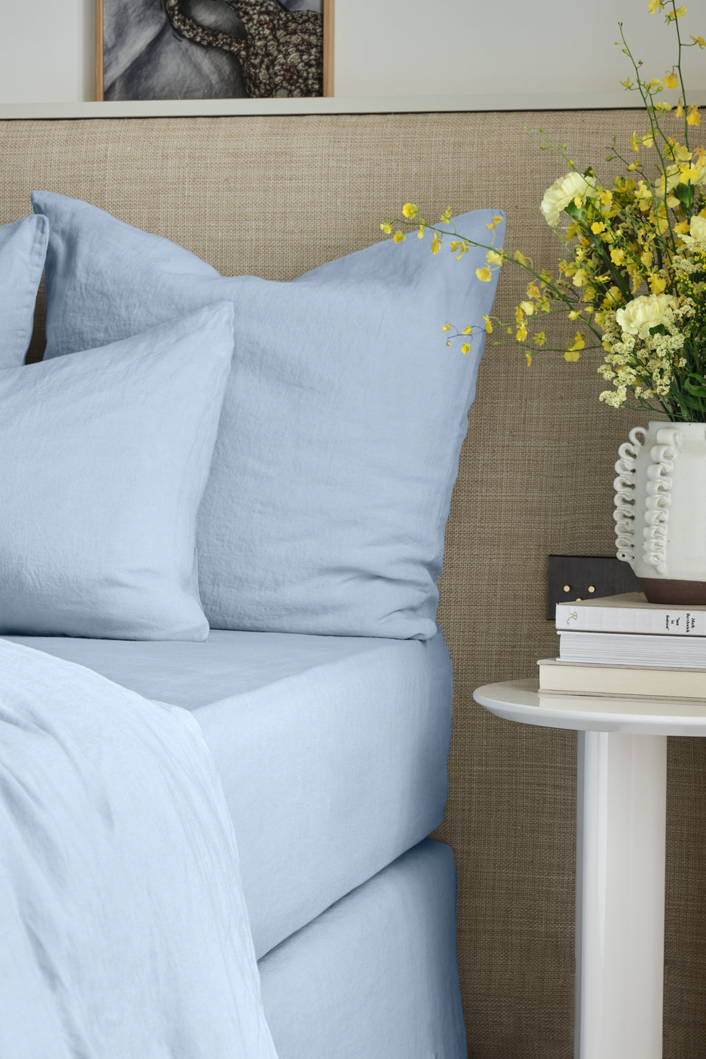 A bed with light blue sheets and pillows beside a beige headboard. A round white nightstand holds a vase of yellow flowers, two books, and a decorative object.