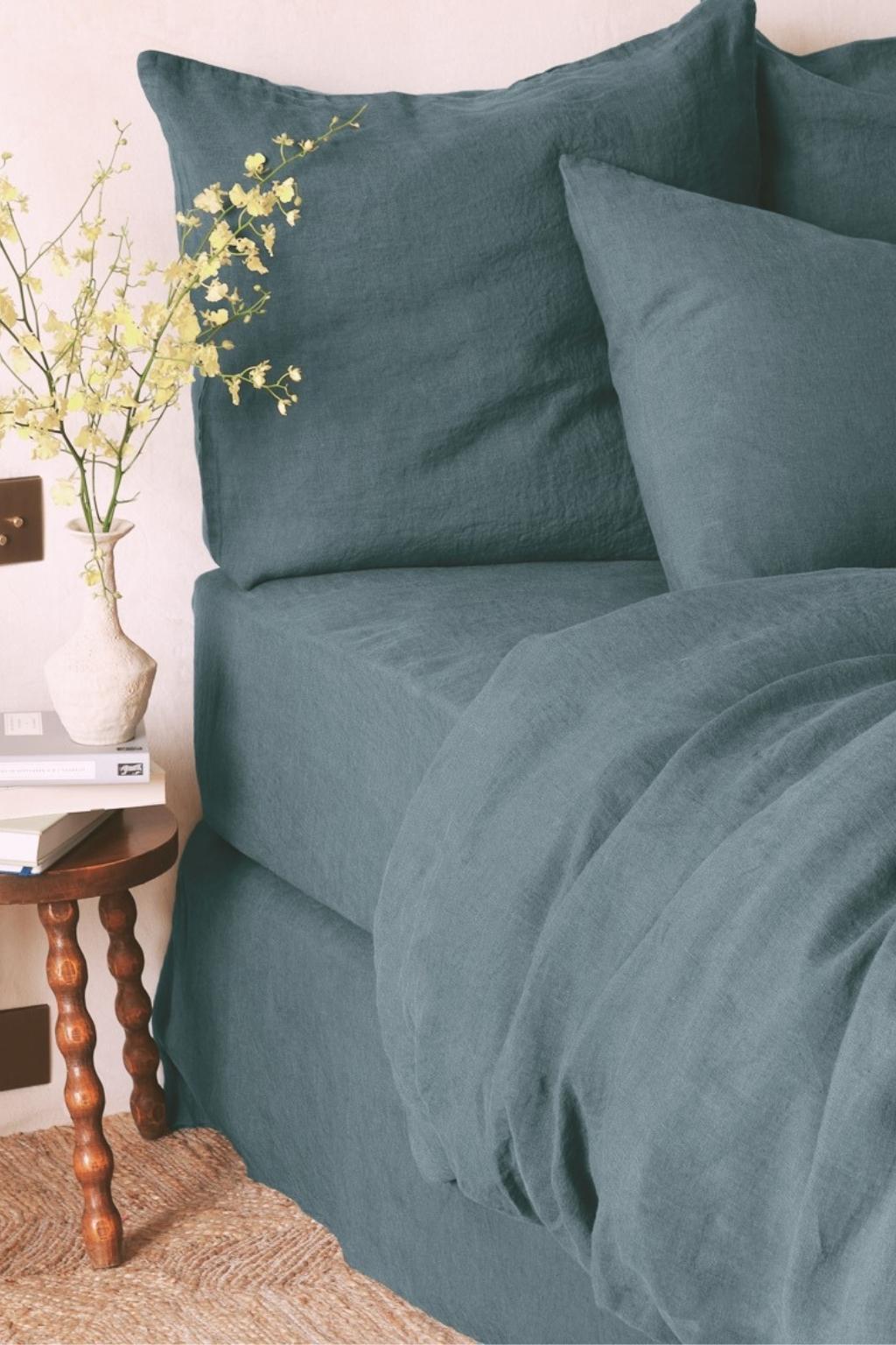 A neatly made bed with blue-green linen bedding sits beside a small wooden table holding a vase of yellow flowers and a stack of books.