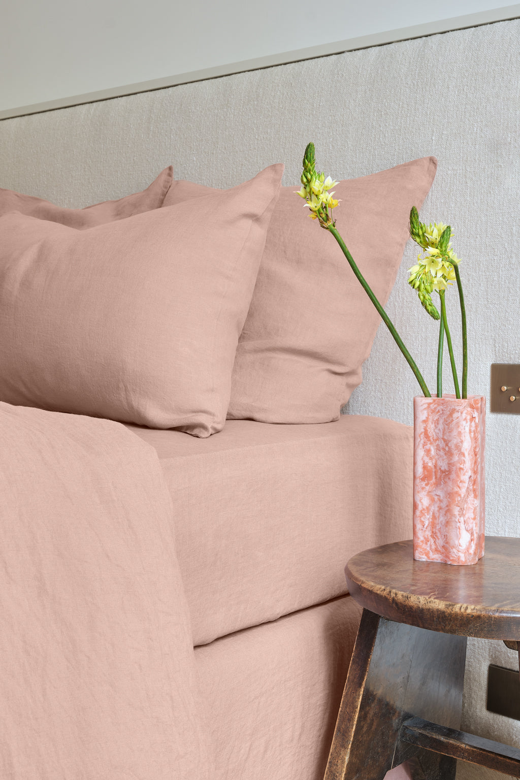 A bed with pale pink linen bedding and pillows is next to a small wooden table holding a pink marbled vase with three white and yellow flowers.