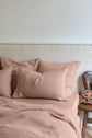 A bed with blush pink linen sheets and pillows is next to a rustic wooden chair holding a small leather bag against a light-colored wall.