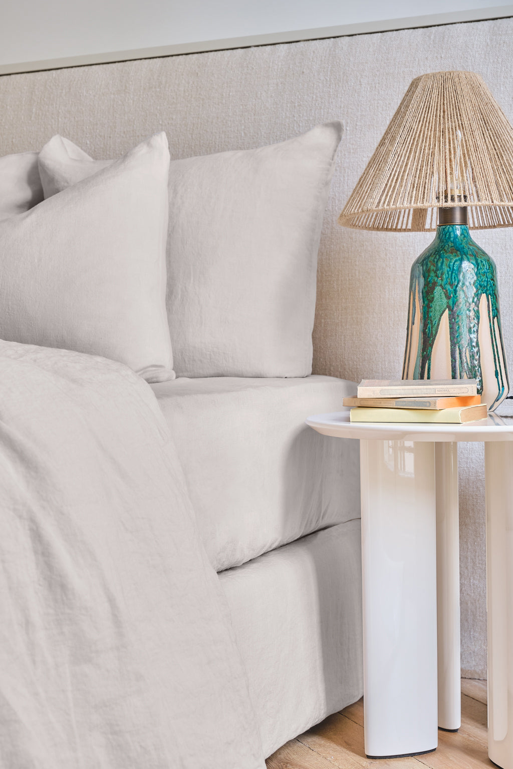 A neatly made bed with light beige linens beside a white nightstand holding books and a decorative lamp with a turquoise and white ceramic base and a woven shade.