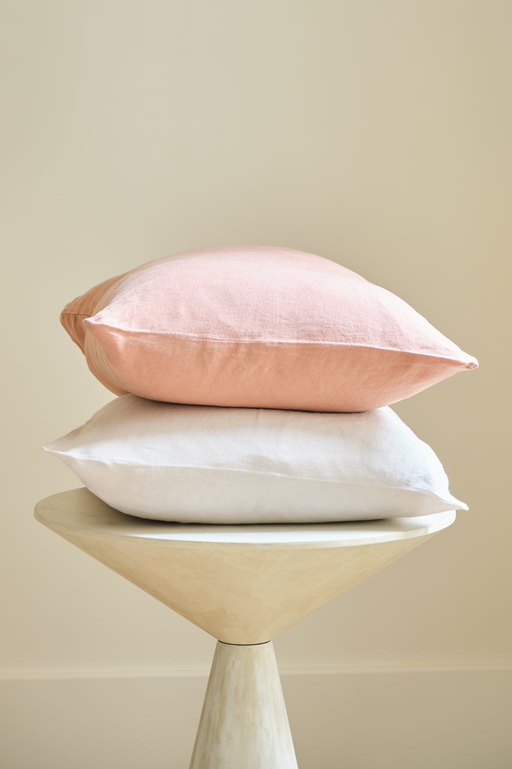 Two pillows, one pink and one white, are stacked neatly on a small, round, light-colored wooden stool against a neutral, beige background.