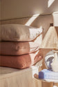 Three stacked pillows in neutral tones sit on a bed next to a table with a lamp and a blue book, all bathed in warm sunlight.
