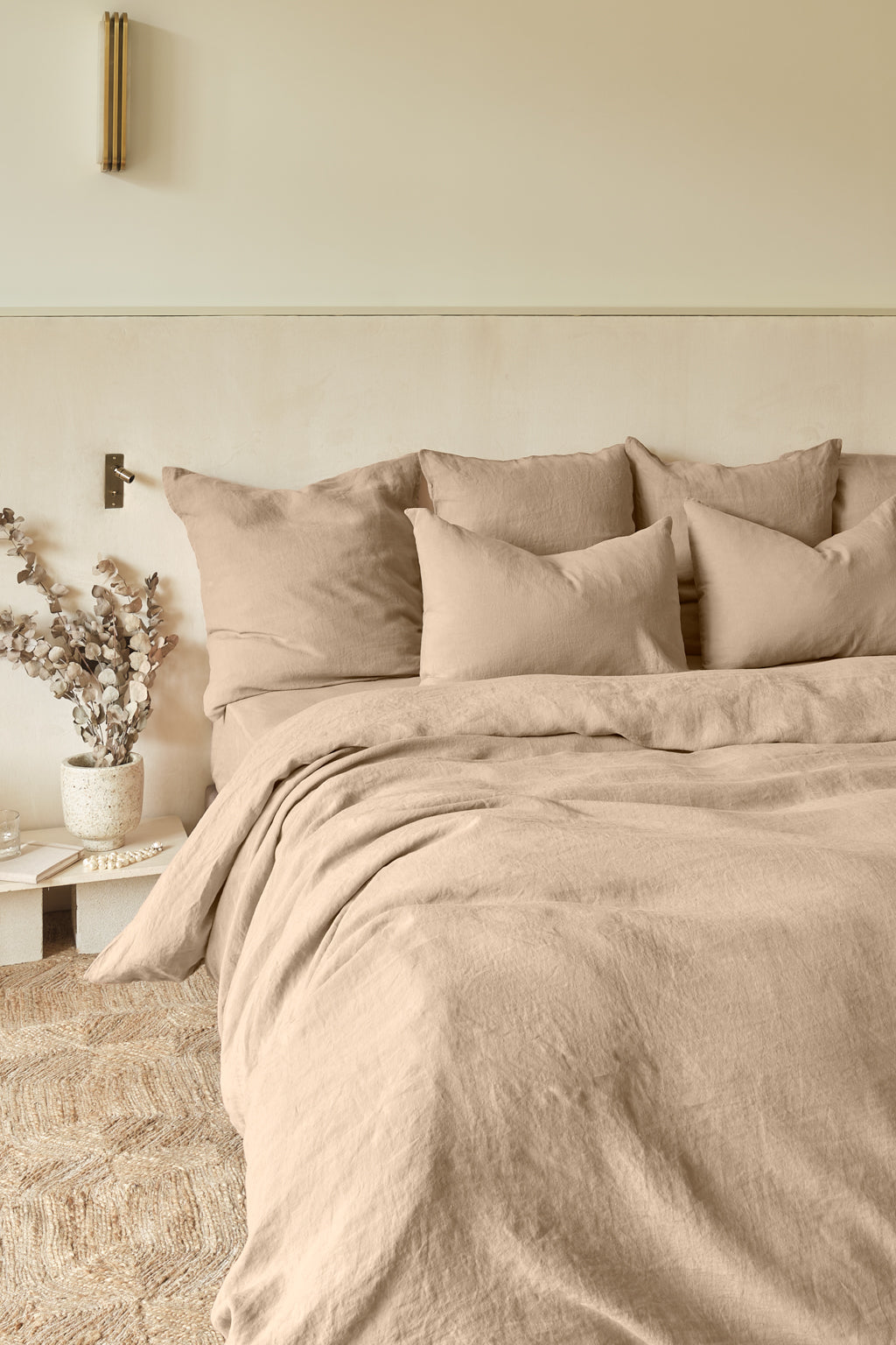 A neatly made bed with beige linen bedding and multiple pillows against a light wall, beside a white nightstand with a potted plant and a brass wall sconce above.