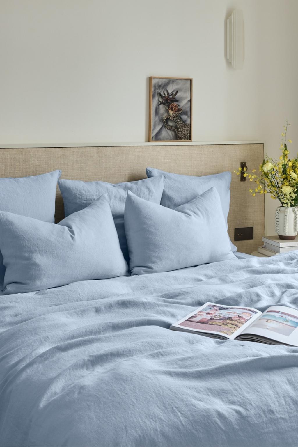 A neatly made bed with light blue bedding and pillows, a nightstand with a vase of yellow flowers, an open magazine, and a framed bird artwork on the wall above the bed.
