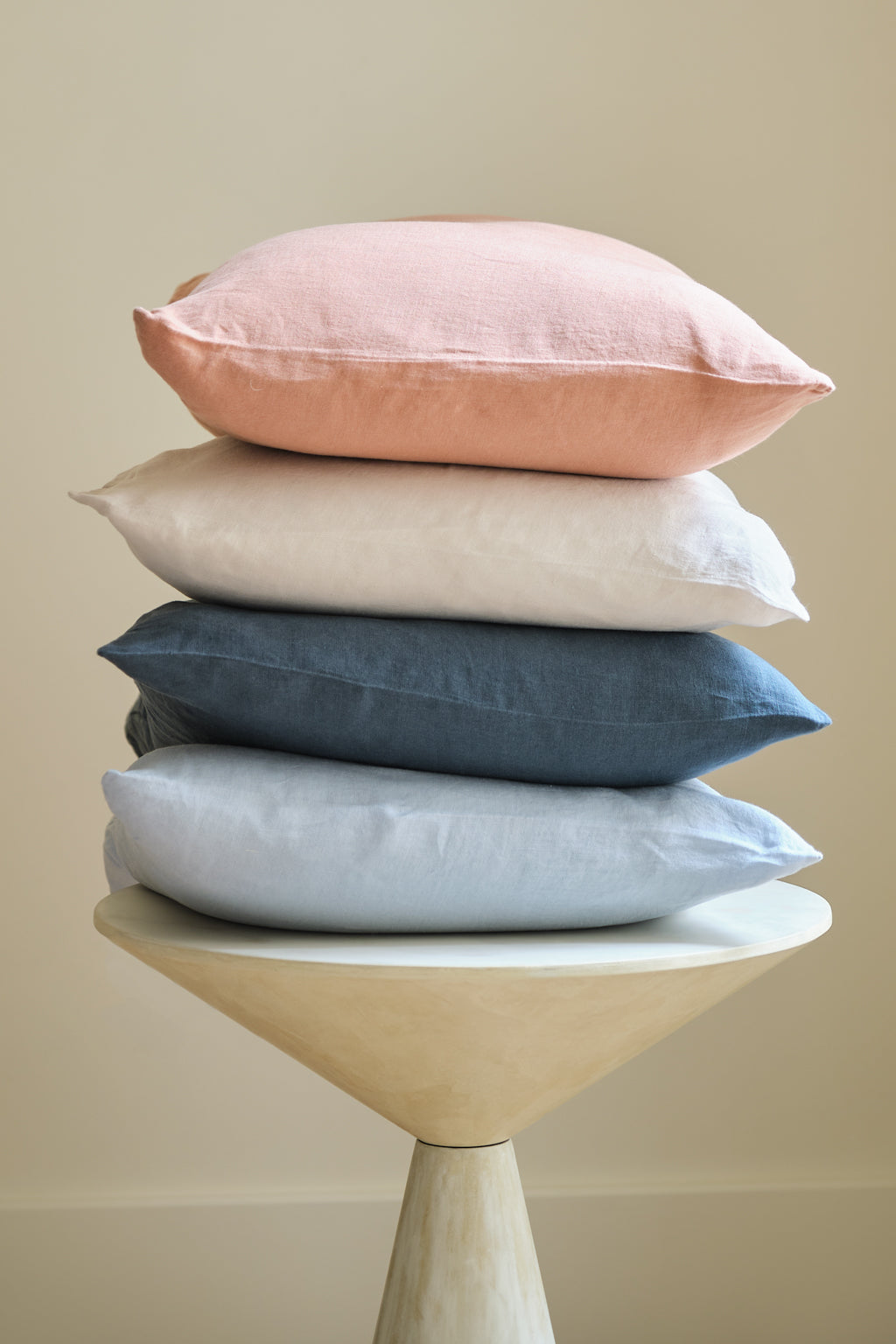 Four pillows in pastel pink, white, blue-gray, and light blue are stacked on a round, modern beige stool against a plain, light neutral background.
