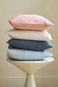 Four pillows in pastel pink, white, blue-gray, and light blue are stacked on a round, modern beige stool against a plain, light neutral background.