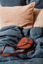 A brown leather crossbody bag rests on a blue blanket, with beige and blue pillows in the background on a neatly made bed.