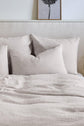 A neatly made bed with three large, beige pillows and a matching comforter, set against a light headboard. A framed black and white flower print hangs on the wall above the bed.