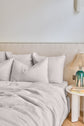 A neatly made bed with white linens and large pillows is next to a round white nightstand holding a lamp with a woven shade, a green base, and a stack of books. The background features a beige fabric headboard.