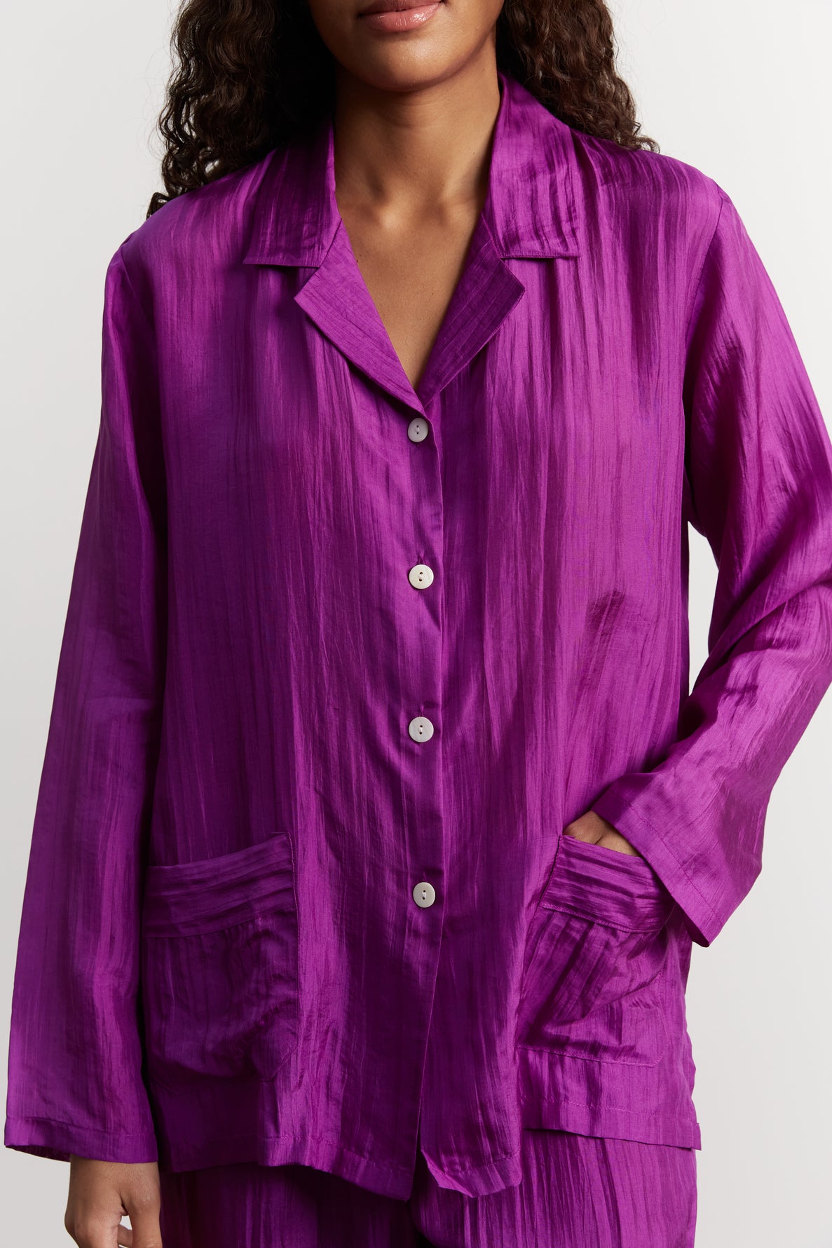 A person wearing a loose, shiny purple button-up shirt with collar and two front pockets, reminiscent of a chic pyjama en soie. The person has one hand in a pocket and their face is partially out of frame.