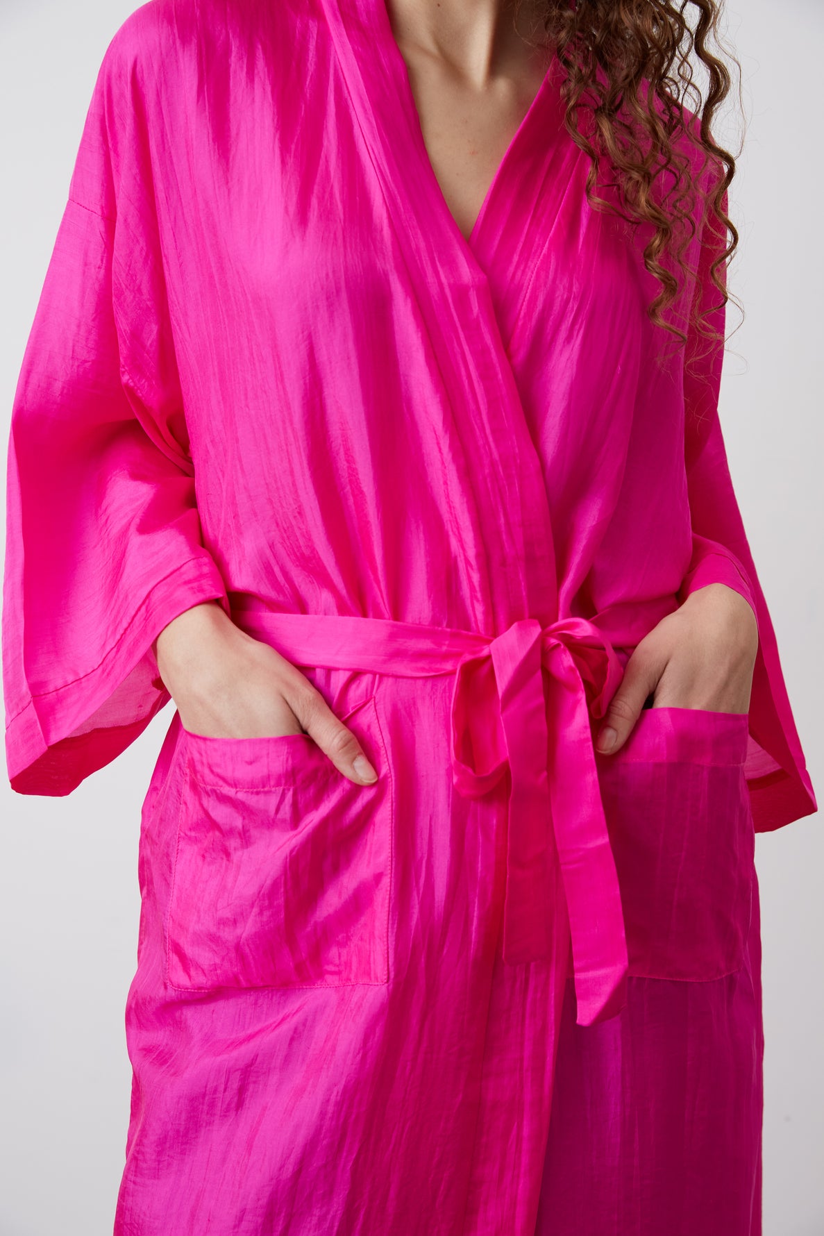 A person wearing a bright pink, belted robe with a ceinture amovible stands with their hands in the robe’s front pockets. The image is cropped to show only the torso and part of the arm, with long curly hair partially visible.