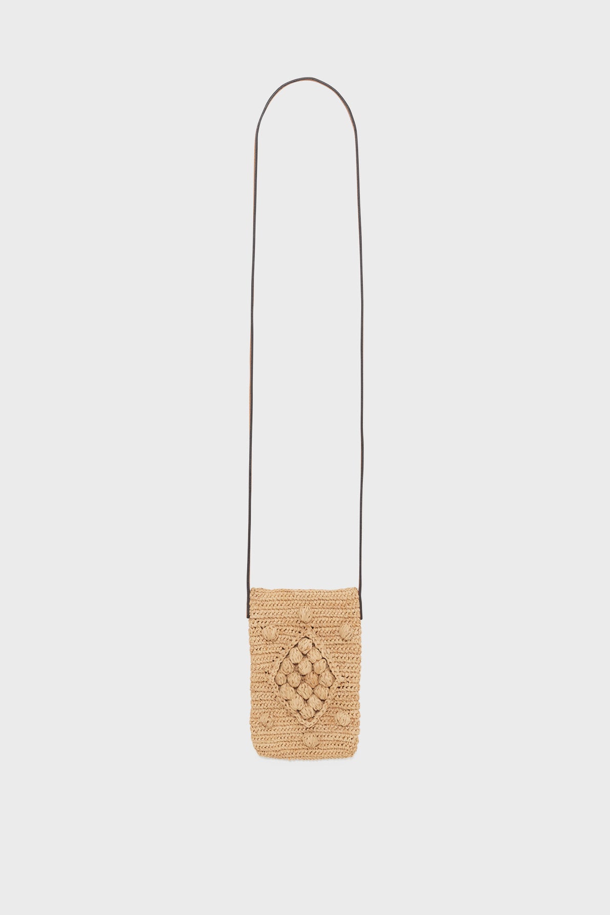 A small, rectangular woven straw crossbody bag crafted through travail artisanal, featuring a long, thin dark bandoulière cuir strap and a decorative diamond pattern on the front, displayed against a plain white background.