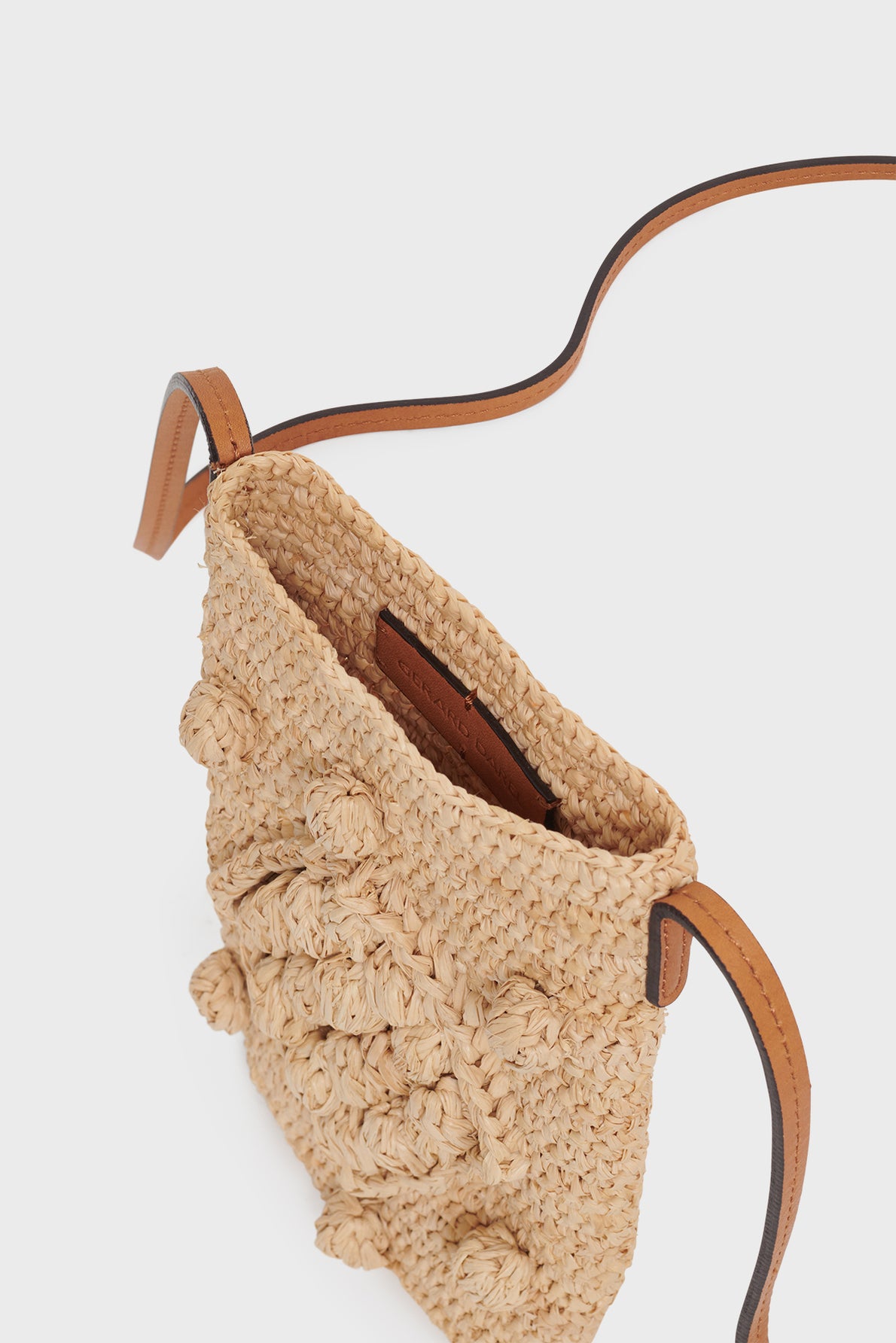 A close-up of a woven straw crossbody bag, crafted with travail artisanal, featuring a thin bandoulière cuir and textured detailing on the front, shown from an overhead angle.