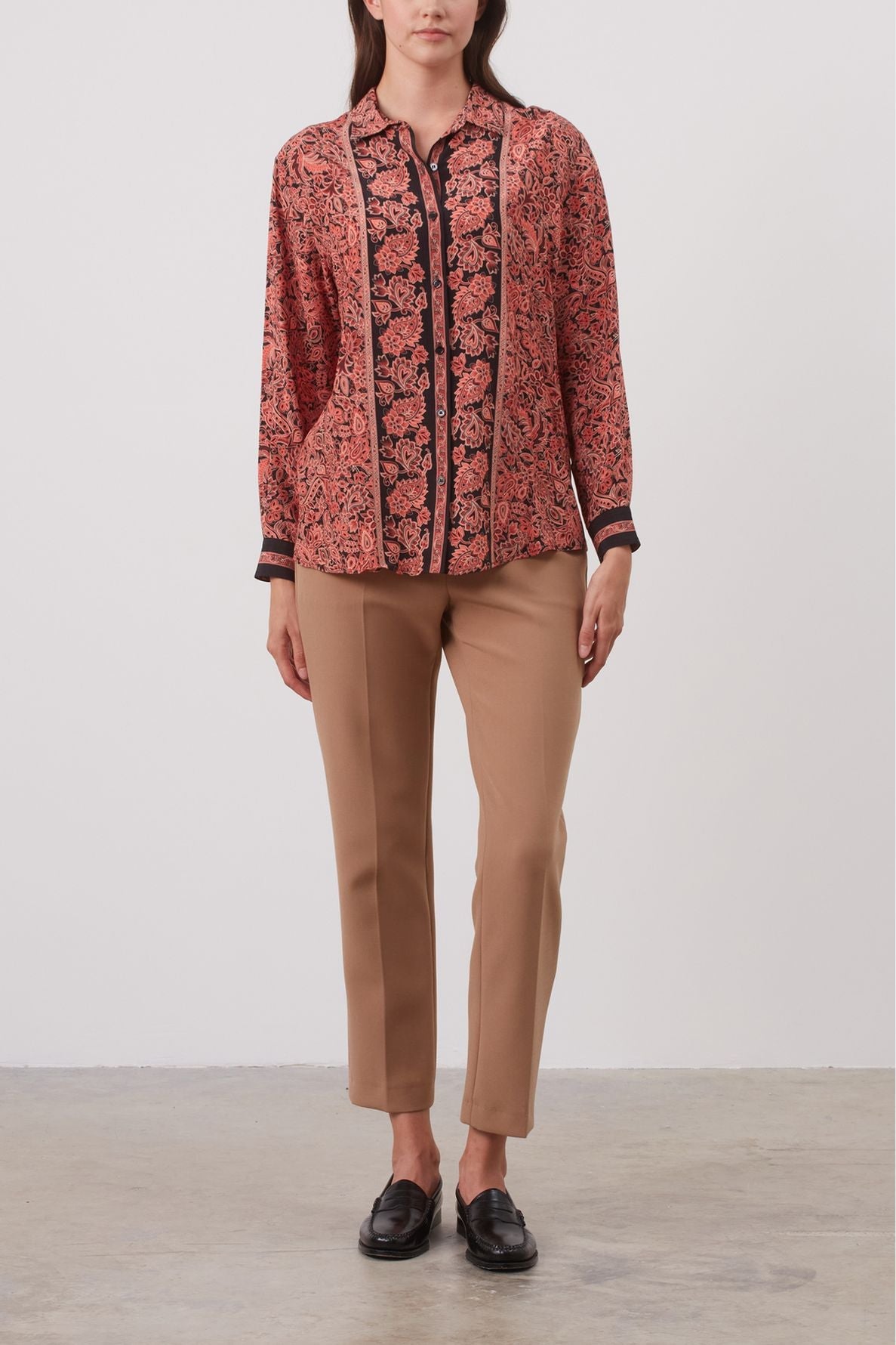 A woman stands against a plain background wearing a red and black floral blouse, tan straight-leg pants, and black loafers. Her hands are relaxed by her sides.