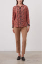 A woman stands against a plain background wearing a red and black floral blouse, tan straight-leg pants, and black loafers. Her hands are relaxed by her sides.