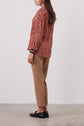 A person stands sideways wearing a patterned red long-sleeve blouse, tan ankle-length pants, and black loafers, against a plain light background.