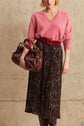 A person wears a pink V-neck sweater, a mousseline plissée jupe midi with a motif papillon and red belt, holds a brown handbag, and stands against a neutral textured wall.