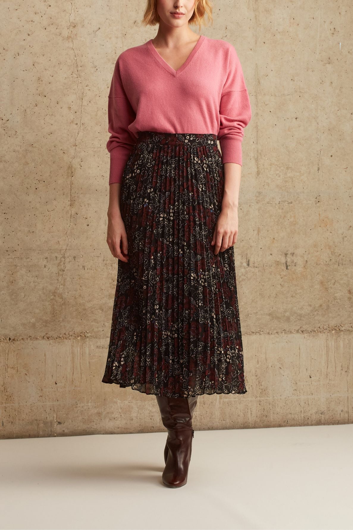 A woman stands against a beige textured wall, wearing a pink V-neck sweater, a dark jupe midi in mousseline plissée with motif papillon, and brown knee-high boots. Her hands rest by her sides. Her face is partially out of frame.