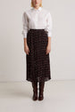 A person stands against a plain background, wearing a white button-down shirt, a black jupe midi plissée with a subtle motif fleuri, and dark brown knee-high boots.
