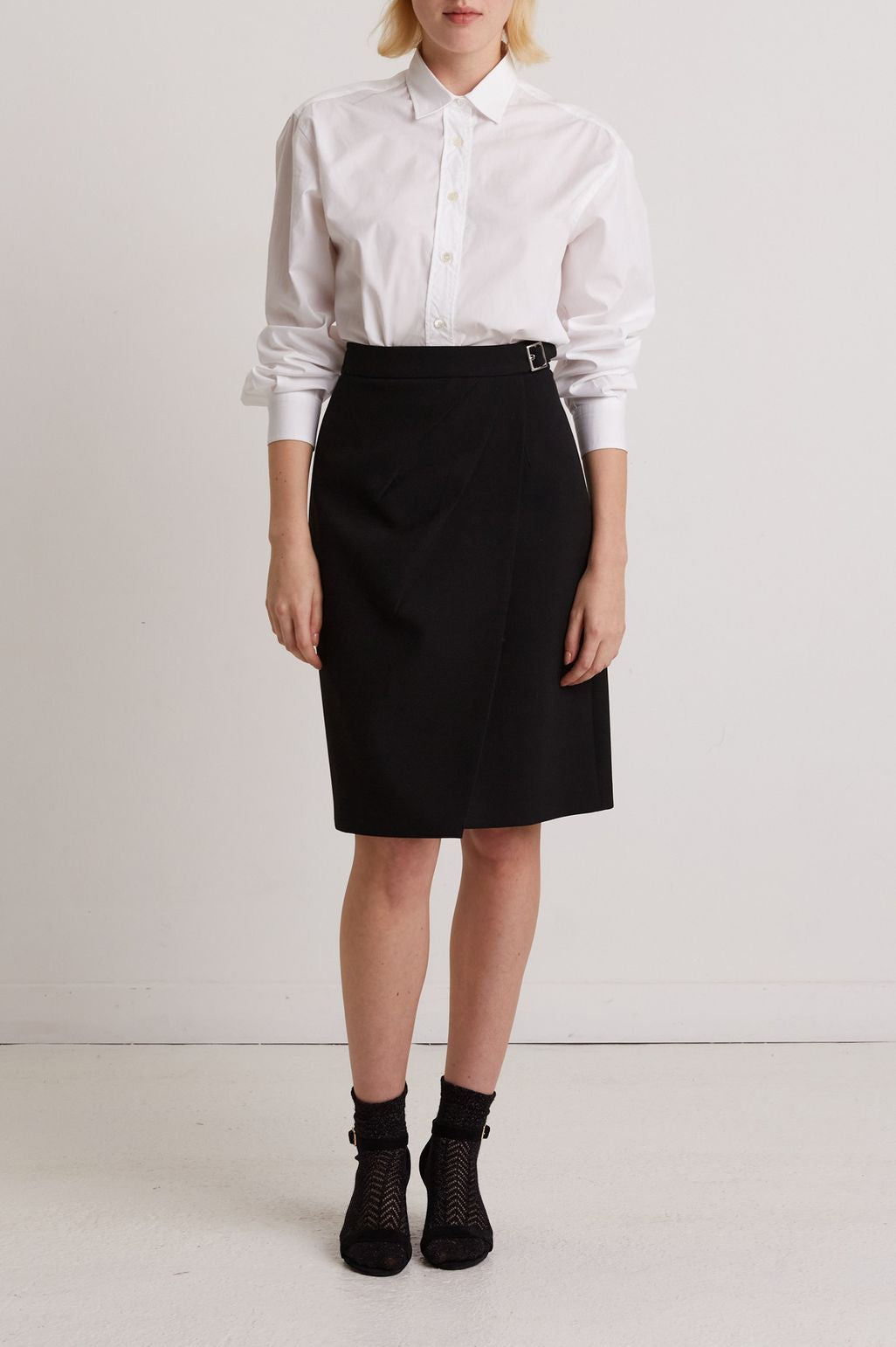 Person standing against a white wall wearing a white long-sleeve button-up shirt tucked into a black knee-length skirt, with black socks and black shoes. The face is not visible in the image.
