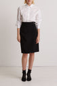 Person standing against a white wall wearing a white long-sleeve button-up shirt tucked into a black knee-length skirt, with black socks and black shoes. The face is not visible in the image.