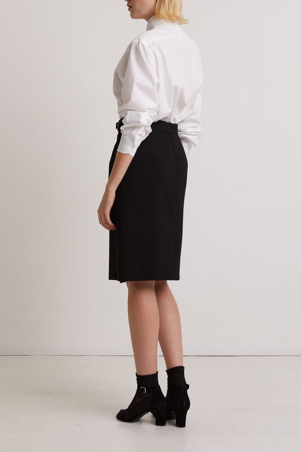 A person stands sideways, wearing a white collared shirt tucked into a black knee-length skirt, black ankle socks, and black heeled shoes. The background is plain and white.