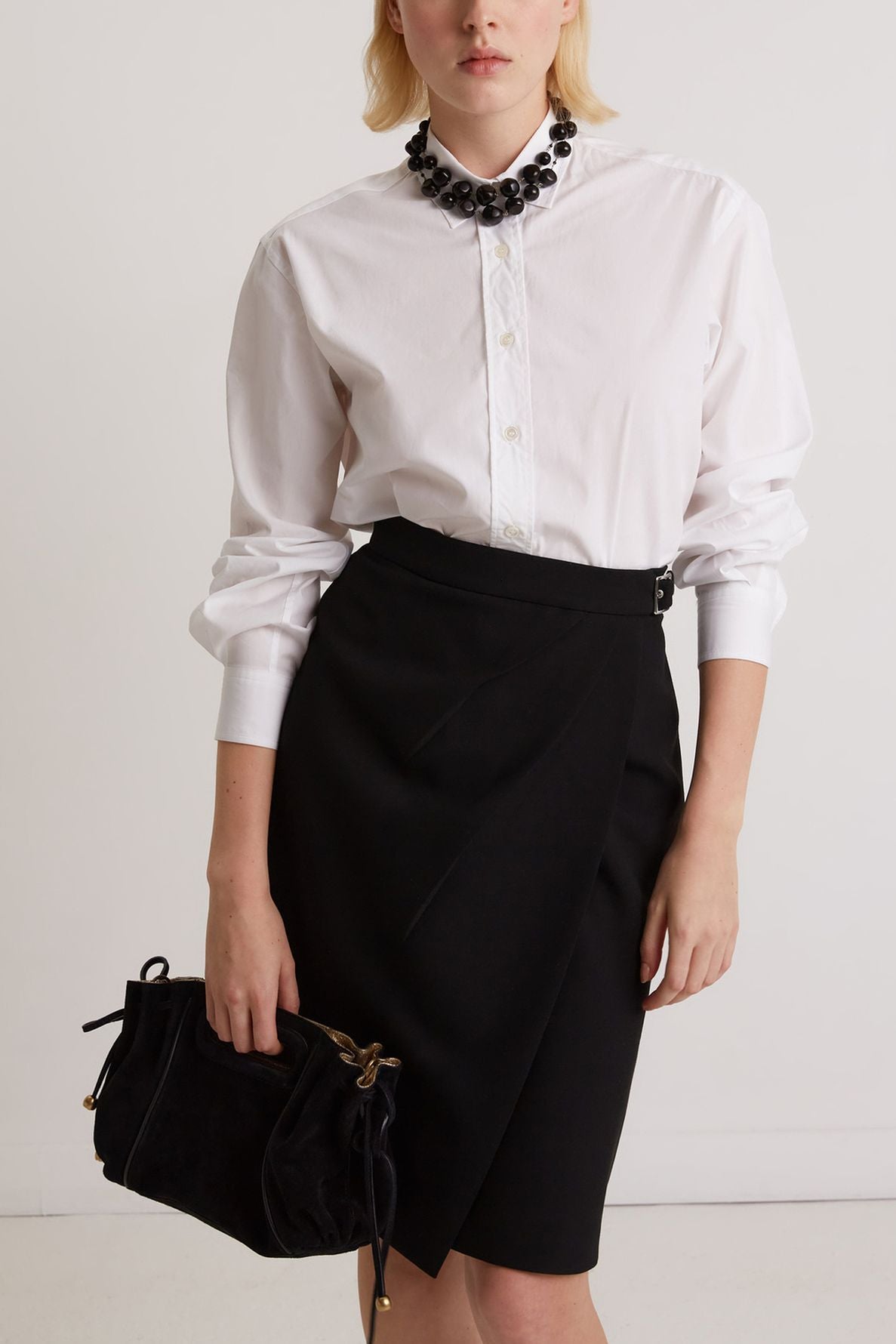 A person wearing a white button-up shirt, black wrap skirt, statement necklace, and holding a black handbag, standing against a plain white background.