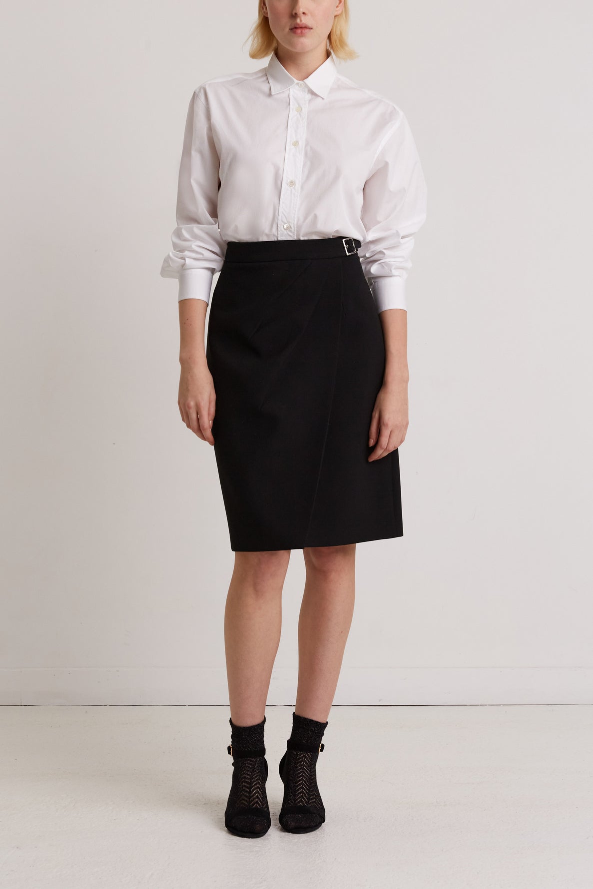 A person stands against a plain white wall, wearing a white button-up shirt, a black knee-length wrap skirt, and black ankle socks with black shoes. Their arms are relaxed by their sides.