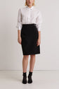A person stands against a plain white wall, wearing a white button-up shirt, a black knee-length skirt, black socks, and black shoes. Their hands are relaxed by their sides.