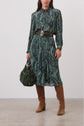 A woman wears a long-sleeved green patterned dress with a wide black belt, carries a green handbag, and pairs the outfit with knee-high brown suede boots.