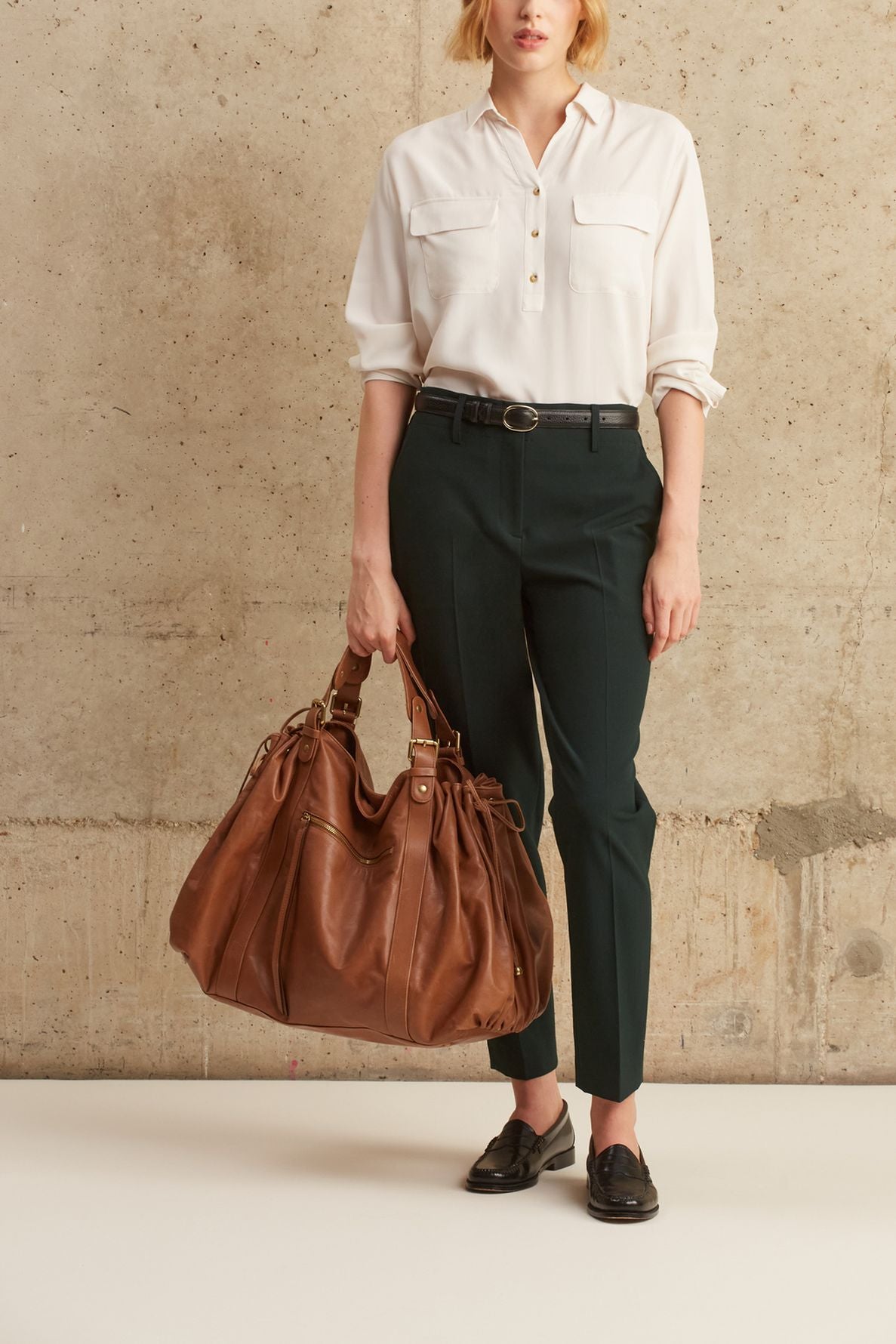 Person in a white button-up shirt, dark green trousers, and black loafers stands against a concrete wall, holding a large brown leather bag in one hand. The photo is cropped above the persons eyes.