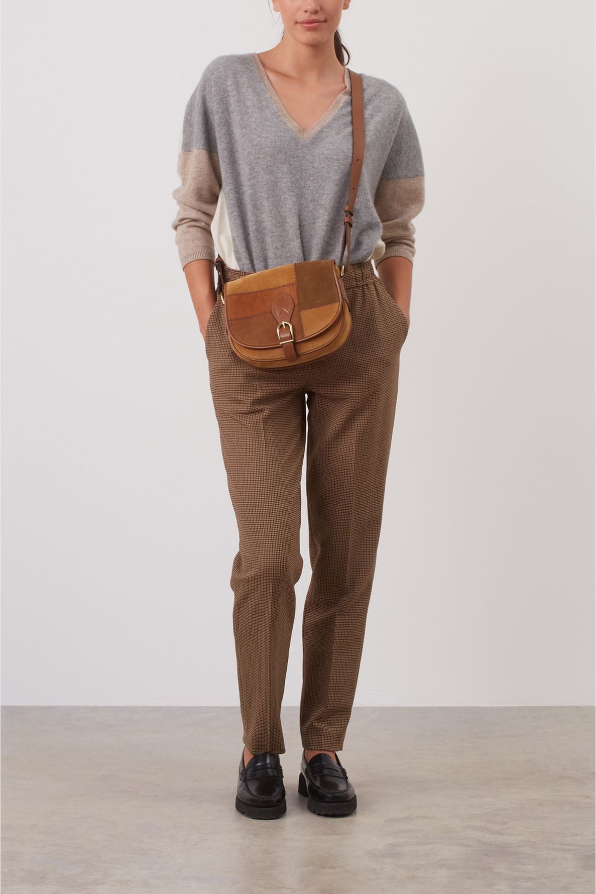 Woman standing against a blank wall, wearing a gray V-neck sweater, brown checked trousers, black shoes, and a brown crossbody bag. Her hands are in her pockets.
