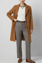 Person wearing a long tan coat over a white top, gray pantalon prince-de-galles coupe droite 7/8, brown belt, and brown loafers, accessorized with a necklace, standing against a plain light background.