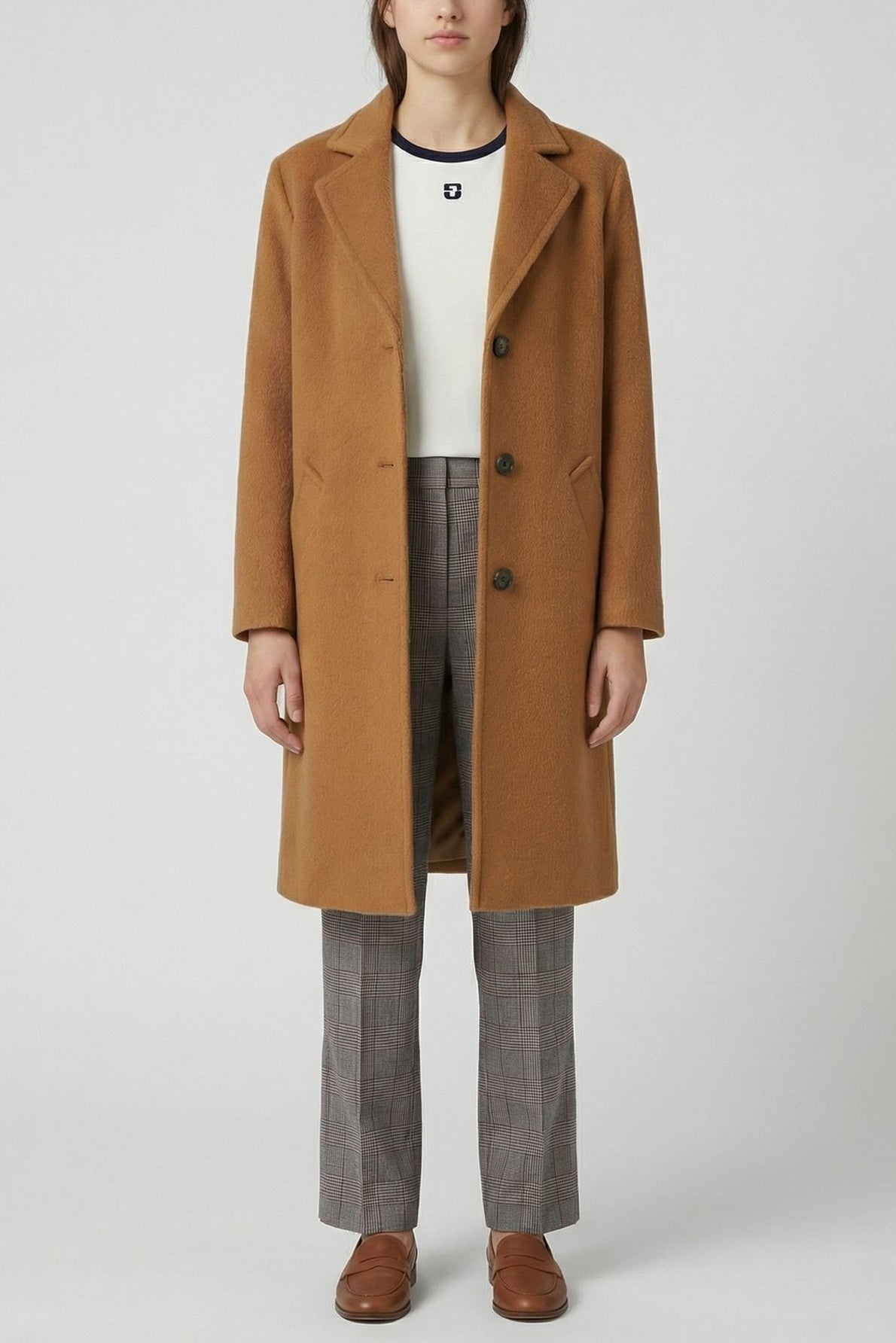 A person wearing a long camel coat over a white top and coupe droite pantalon prince-de-galles 7/8, paired with brown loafers, standing against a plain light background.