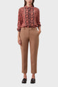 A person wearing a red and black patterned blouse tucked into tan tailored pants with a belt, hands in pockets, and black loafers, standing against a plain white background.