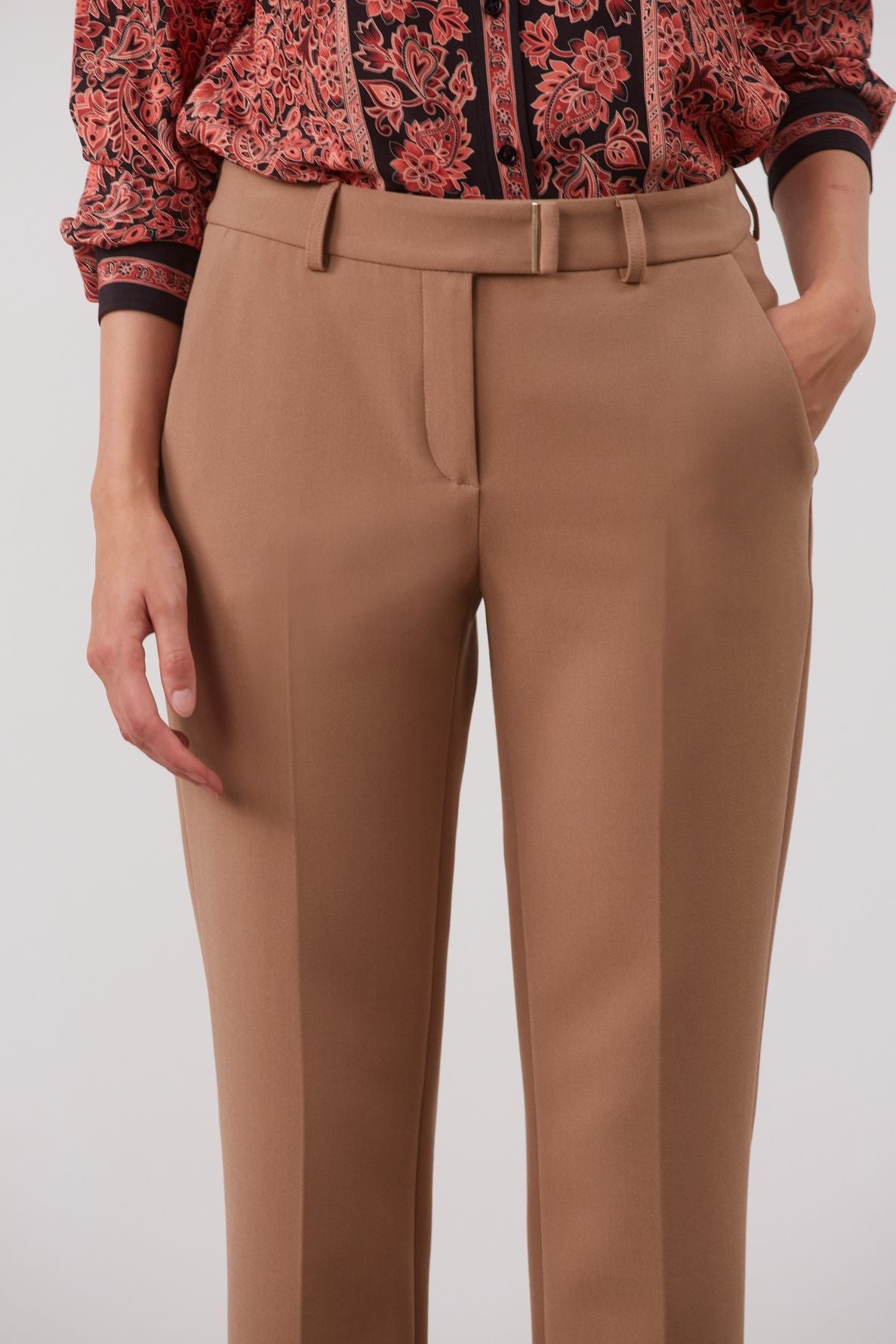A person wears tan dress pants with hands in the pockets and a patterned red and black long-sleeve shirt tucked in. The upper body is partially visible against a plain background.