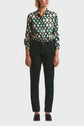 A person stands wearing a green and cream geometric-patterned button-up shirt, dark green pantalon cigarette en velours côtelé, and black shoes. Their hands are at their sides, and their face is not visible.