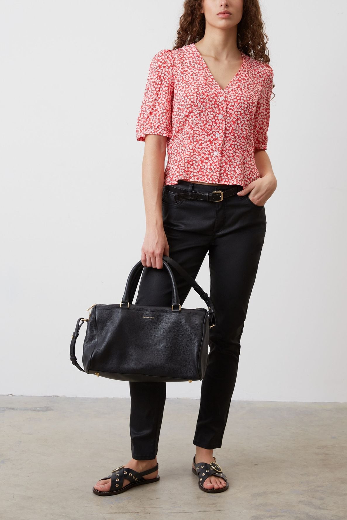 A woman wearing a red patterned blouse, black pants, and black sandals stands holding a large black handbag. She has long curly hair and is standing against a plain white wall.