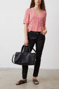 A woman wearing a red patterned blouse, black pants, and black sandals stands holding a large black handbag. She has long curly hair and is standing against a plain white wall.