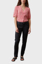 A person stands wearing a red and white patterned blouse, black pants, and black sandals against a plain light background. Their arms are relaxed at their sides.
