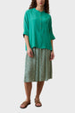 A woman wearing a loose oversize green blouse with buttons, a green floral knee-length skirt, and brown sandals poses against a plain light background.