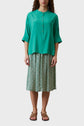 A woman wears an oversize teal, button-up, three-quarter sleeve blouse and a green, floral midi skirt with brown sandals. She stands facing forward against a plain background.