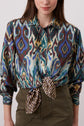 A woman wearing a colorful patterned button-up shirt tied at the waist and olive green high-waisted pants, standing against a plain background.