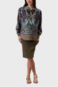 A woman stands wearing a colorful patterned blouse, an olive knee-length skirt, and gold sandals. She is smiling with one hand in her pocket against a plain light background.