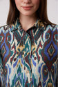 A woman with long brown hair wearing a colorful patterned shirt with a collar and buttons, smiling slightly. The shirt features abstract shapes in teal, blue, yellow, maroon, and cream.
