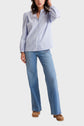 A person wearing a light blue striped button-up shirt, blue flared jeans, and brown clogs stands with one hand touching their hair against a plain light background.