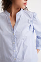 Woman wearing a light blue, pinstriped button-up shirt with a collarless neckline, standing against a plain white background and facing slightly to the side.