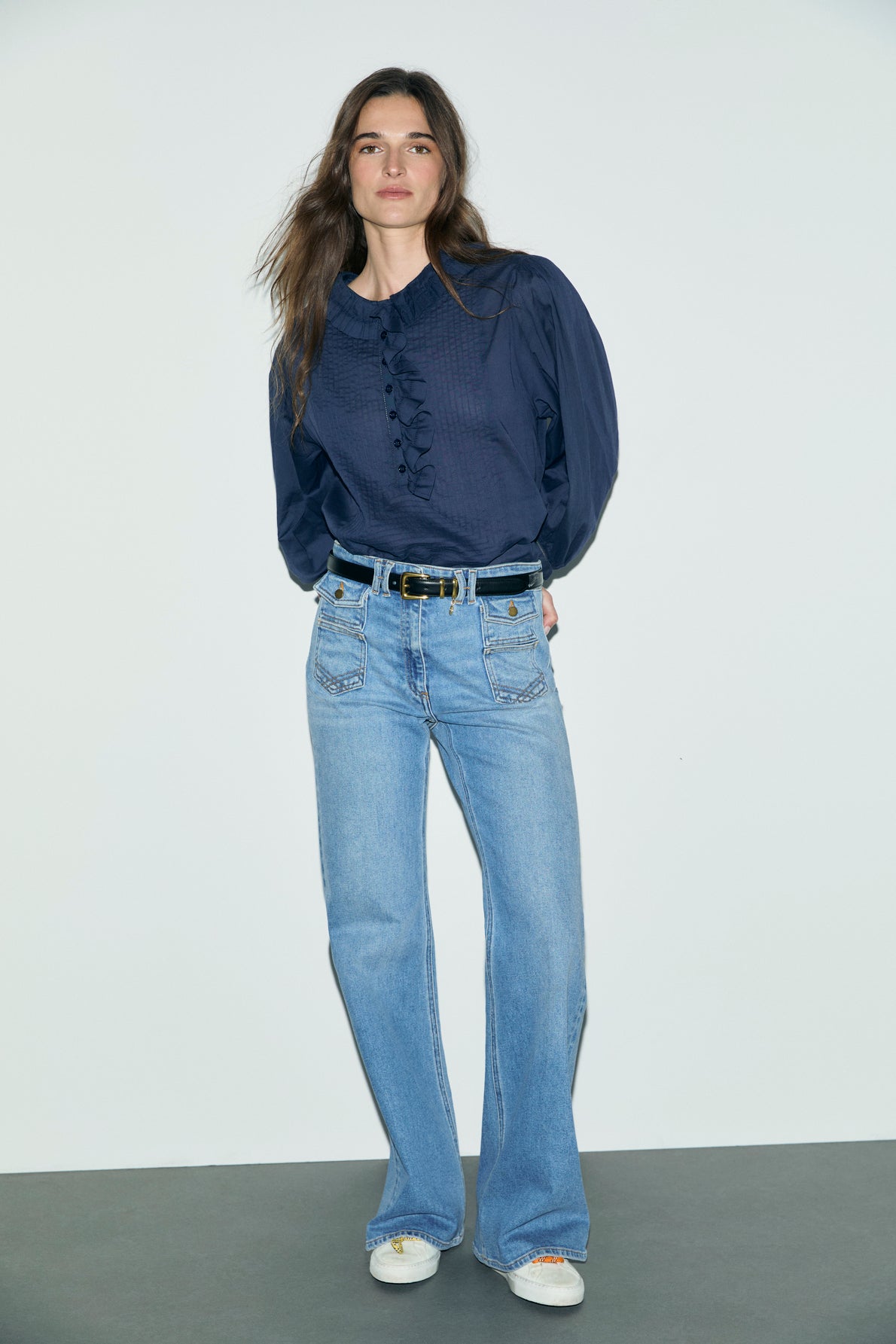 A person stands against a plain background wearing a navy blouse with textured buttons, light blue pantalon long taille haute jeans, a black belt, and white sneakers. Their hands are behind their back and they have long, wavy hair.